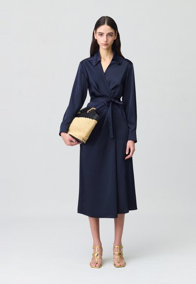 Vestido envolvente de satén en color marino con mangas largas y cinturón en la cintura, acompañado de un bolso negro de paja y sandalias doradas con tiras.