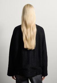 Maglione oversize nero testurizzato con maniche lunghe e orlo arrotondato, con dettagli a bottone sul bordo inferiore. Si vede una lunga chioma bionda e liscia.