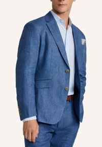 Mann trägt einen blauen Leinenanzug mit braunen Knöpfen über einem hellblauen, gestreiften Hemd, braunem Gürtel und gemustertem Einstecktuch.