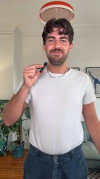 Melns mikrofons, ko tur cilvēks baltā T-kreklā un zilās džinsās, ar īsiem cirtainiem matiem un sudraba ķēdi, gaišā telpā.