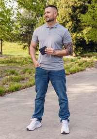 Gray polo shirt, blue jeans, and white sneakers. Holding a black wallet, standing in a park with greenery and a path in the background.