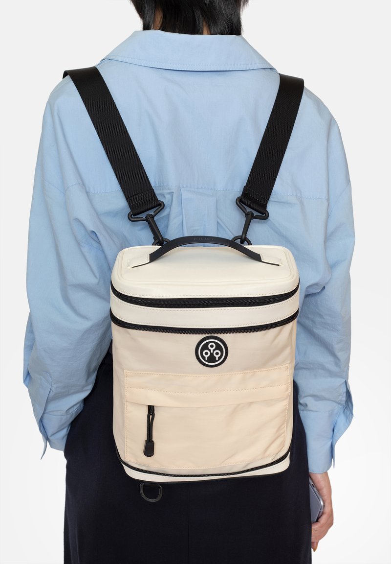 Sac à dos avec un corps crème, des accents noirs et une poignée supérieure. Il dispose d'un compartiment zippé et d'une poche avant avec un logo noir.