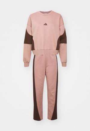 Tuta di colore rosa e marrone realizzata in tessuto morbido. La parte superiore presenta un collo rotondo e maniche a blocchi di colore, mentre i pantaloni hanno accenti laterali.