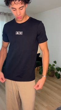 Joven con cabello rizado, vestido con camiseta negra y pantalones beige, en el interior junto a una planta y cortinas de ventana, mirando hacia abajo.