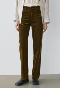 Person iført brune kordfløjlsbukser med lige ben, stribet lysebeige skjorte og sorte sko med firkantet tå, stående på grå gulv.