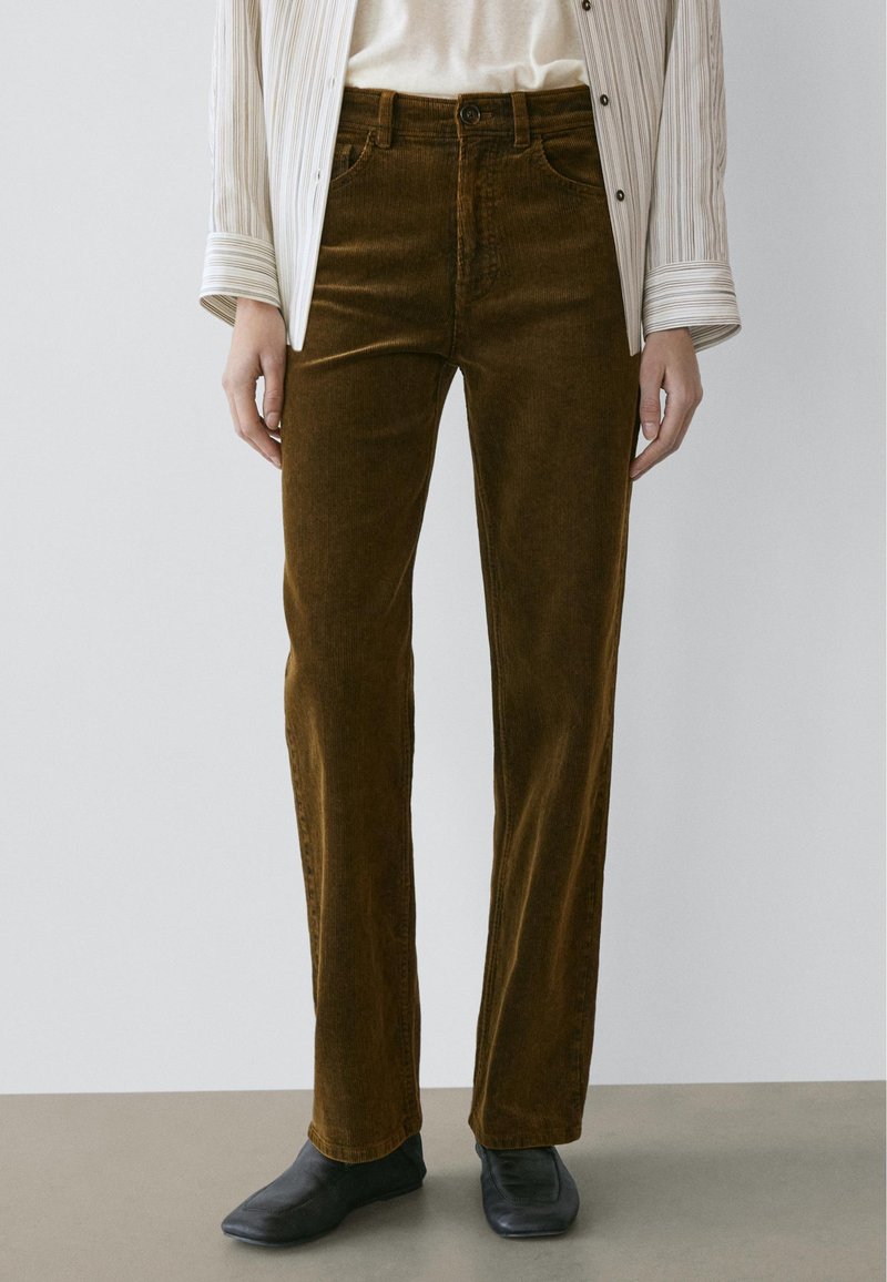 Person iført brune kordfløjlsbukser med lige ben, stribet lysebeige skjorte og sorte sko med firkantet tå, stående på grå gulv.