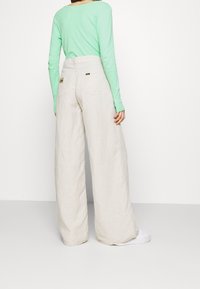 Svetle zelený dlouhý rukáv top spárovaný s volnými béžovými lněnými kalhotami se širokým pasem a zadními kapsami. Viditelný detail textury tkaniny.