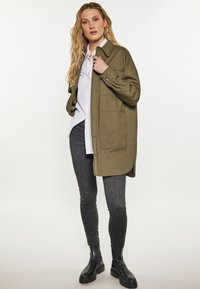 Oliven grøn skjortejakke med løs pasform, store frontlommer, knaplukning, lagdelt over en hvid skjorte og parret med mørke stramme jeans og sorte støvler.