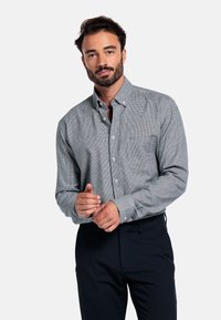 Man met donker haar en baard die de manchetten van een lichtgrijs geruit overhemd aanpast, gekleed in donkere broek, staande tegen een witte achtergrond.