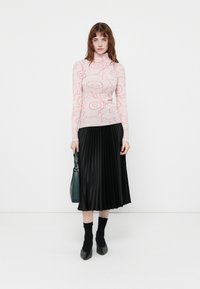 Svijetlosmeđi turtleneck pulover s crvenim cvjetnim uzorkom, uparen s crnom plisiranom midi suknjom. Dodaci uključuju zelenu torbu i crne cipele.