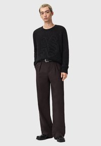 Zwarte gebreide trui met een textuurpatroon, gecombineerd met losse, donkerbruine broeken. Model staat voor een neutrale achtergrond.