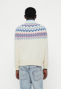 Μπλούζα από πλεκτό κρεμ με μοτίβο Fair Isle σε ανοιχτό μπλε και ροζ. Μανσέτες και τελείωμα με ρεβέρ. Φορεμένο πάνω από ανοιχτό μπλε τζιν παντελόνι.