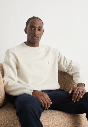 Light beige sweatshirt with a crew neck, ribbed cuffs, small logo detail, worn over dark navy trousers, seated on a textured brown chair.