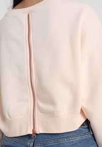 Svetle růžová mikina s krátkým designem, která má viditelný zip na zádech a žebrovaný spodní lem. Měkká textura a uvolněný střih.