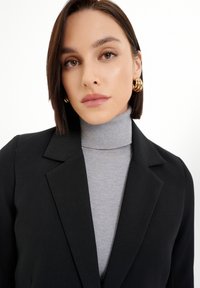 Crni blejzer s oštrim reverima preko prilagođenog sivo turtleneck pulovera. Subjekt nosi velike zlatne prstenaste naušnice. Glatka koža i ravna kosa.