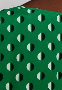 Grüner Stoff mit einem sich wiederholenden Muster aus schwarzen und weißen Halbkreisen. Das Material wirkt glatt mit einem leichten Glanz.
