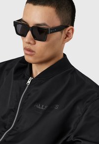 Jeune homme portant des lunettes de soleil rectangulaires noires et une veste bomber noire zippée sur un fond clair uni.