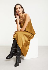Robe en satin moutarde avec une silhouette fluide, ourlet asymétrique, associée à des bottes en cuir noir jusqu'aux genoux et des bijoux en chaîne dorée.