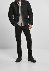 Černá vojenská bunda se zapínáním na zip, předními kapsami, šedá košilová košile, černé džíny a tmavé boty.