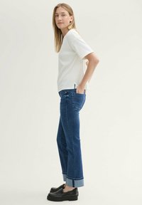 Top corto de manga corta blanco con relieve, jeans de mezclilla azul con puños enrollados y zapatos negros de plataforma. El modelo está de pie con las manos en los bolsillos.