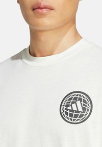 Nahaufnahme einer Person, die ein weißes T-Shirt mit einem schwarzen kreisförmigen Globusdesign und einem Logo mit drei diagonalen Streifen auf der Brust trägt.