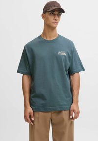 Tricou verde din bumbac, cu mâneci scurte, guler rotund și text imprimat alb "APPAREL STUDIOS" pe pieptul stâng. Șapcă maro.