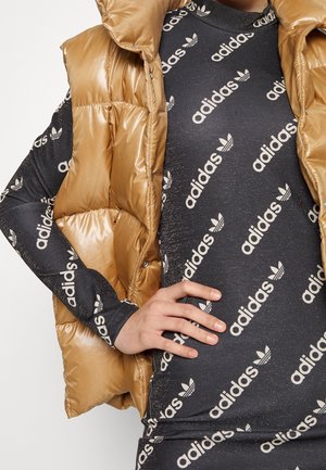Złota kamizelka puchowa na czarnej sukience z długim rękawem, z powtarzającym się białym wzorem logo Adidas. Gładka, błyszcząca tkanina o pikowanej strukturze.