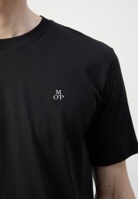 Černé krátké rukávy tričko vyrobené z bavlny, s malým bílým vyšívaným logem na levé části hrudníku. Hladká textura, design s kulatým výstřihem.