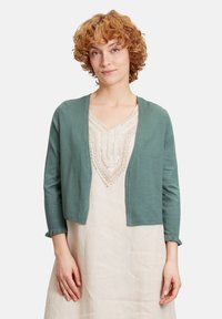 Cardigan leggero color teal, design abbottonato davanti, maniche a tre quarti con polsini arricciati, indossato sopra un vestito beige con ricami intricati.