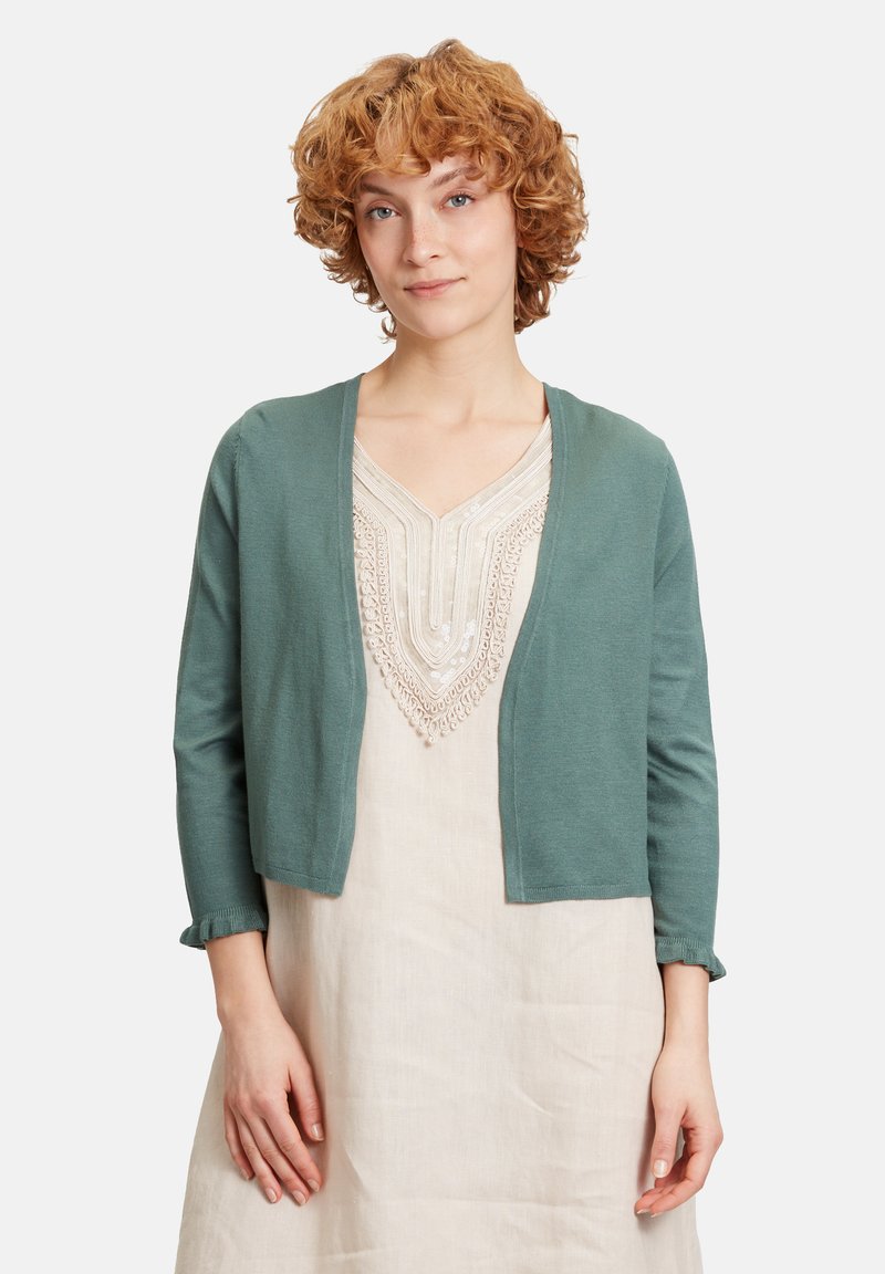 Cardigan leggero color teal, design abbottonato davanti, maniche a tre quarti con polsini arricciati, indossato sopra un vestito beige con ricami intricati.