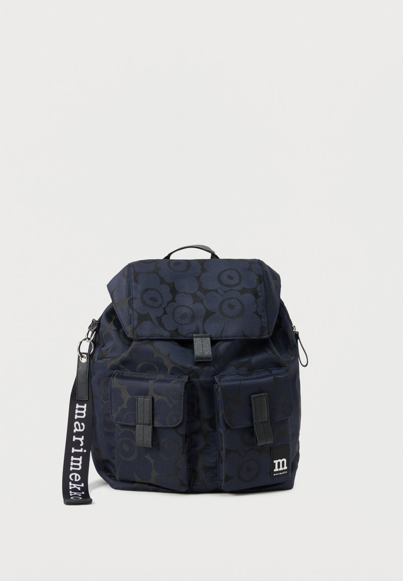 Σκούρο μπλε σακίδιο με λουλουδάτο μοτίβο, δύο εμπρόσθιες τσέπες με καπάκι, χειρολαβή στο πάνω μέρος και μαύρο ιμάντα με την ετικέτα "marimekko" σε λευκό φόντο.