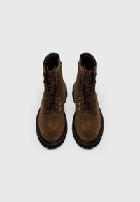 Ruskeat mokkanahasta nyörikengät, joissa on paksu musta kumipohja ja metalliset silmukat, pyöreä kärki ja sivussa vetoketjusuljenta.