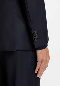 Dunkelblaues Marine-Anzug mit strukturiertem Finish, ausgestattet mit einem geknöpften Manschettenabschluss mit vier schwarzen Knöpfen und kontrastierendem blauem Stoff am Ärmelabschluss.