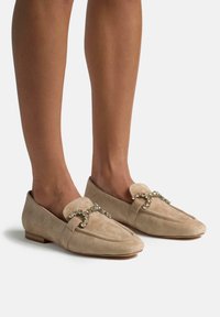 Beige Wildleder Mokassin Slipper mit runder Zehenpartie, ausgestattet mit einem weichen Obermaterial und einem dekorativen, mit Kristallen akzentuierten Riemen über dem Spann.