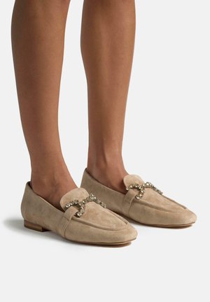 Mocassins en daim beige avec une pointe arrondie, présentant un upper doux et une sangle décorative ornée de cristaux à travers le cou-de-pied.