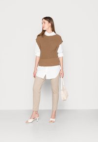 Ruskea neuleliivi valkoisen paidan päällä, yhdistettynä beigen värisiin slim-fit-housuihin ja vaaleisiin korkokenkiin. Pieni tekstuuripintainen käsilaukku.