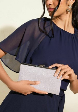Clutch cu glitter argintiu, cu o clapetă metalică, ținut în mână. Persoana poartă o rochie albastră marină cu mâneci transparente. Textură netedă.
