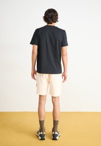 Svart bomull t-skjorte, kremfargede shorts, grå sokker og sneakers med blå detaljer; modell som vender bort fra kameraet mot en lys bakgrunn og gul gulv.