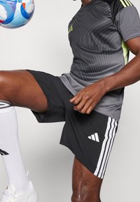 Svarta sportshorts med vita ränder, tillsammans med en grå och svart mönstrad t-shirt med gröna detaljer. En fotboll i närheten.