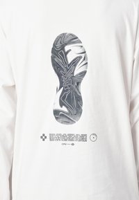 Fehér, hosszú ujjú ing, amelyen egy fekete grafika látható cipőtalpról és alatta részletes szöveg. Simára megmunkált pamut anyag és kerek nyakú dizájn.
