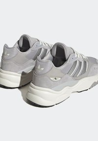 Ljusgrå Adidas-sneakers med vita snören, tre reflektiva sidosträck, vadderad häl och tjocka vita sulor sedda från bak- och sidor.