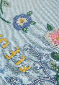 Blue denim fabric with embroidered flowers in purple, pink, and green, alongside yellow embroidered text reading "FLY."