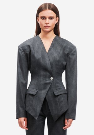 Donna in piedi, rivolta verso l'obiettivo, indossa un blazer grigio a righe sottili con spalle imbottite esagerate e pantaloni abbinati su uno sfondo bianco.