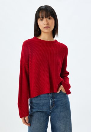 Roter gerippter Strickpullover mit rundem Ausschnitt und langen Ärmeln, im cropped Design, kombiniert mit hochgeschnittenen blauen Jeans.
