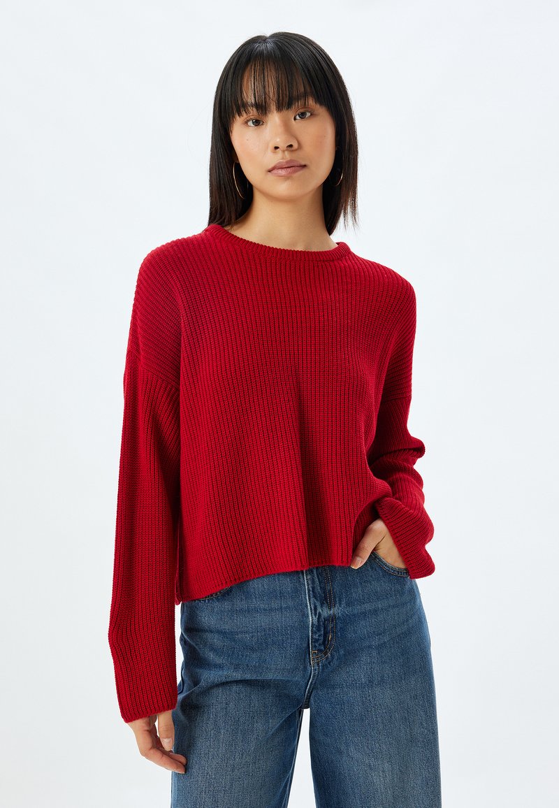 Roter gerippter Strickpullover mit rundem Ausschnitt und langen Ärmeln, im cropped Design, kombiniert mit hochgeschnittenen blauen Jeans.