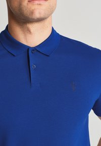Sininen polo-paita, joka on valmistettu teksturoidusta kankaasta, sisältää klassisen kauluksen, kaksipainikkeisen avauksen ja pienen, tumman logon vasemmassa rinnassa.