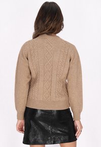 Beige gestrickter Pullover mit einem Kabelmuster auf der Rückseite und geripptem Bund, kombiniert mit einem schwarzen glänzenden Minirock.