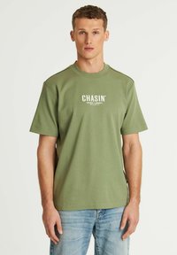 Alyvos žalios spalvos trumpų rankovių marškinėliai, pagaminti iš medvilnės, su apvaliu kaklu ir baltu užrašu "CHASIN'" priekyje. Paprastas dizainas.