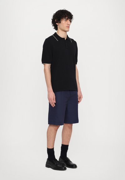 Polo shirt noir à côtes avec col contrasté, associé à des shorts navy. Le mannequin porte des mocassins noirs et des chaussettes foncées. Tenue simple et décontractée.