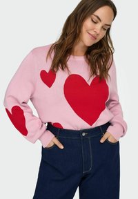 Růžový pletený svetr zdobený velkými červenými srdci, s žebrovanými manžetami a kulatým výstřihem, kombinovaný s tmavými džínami.
